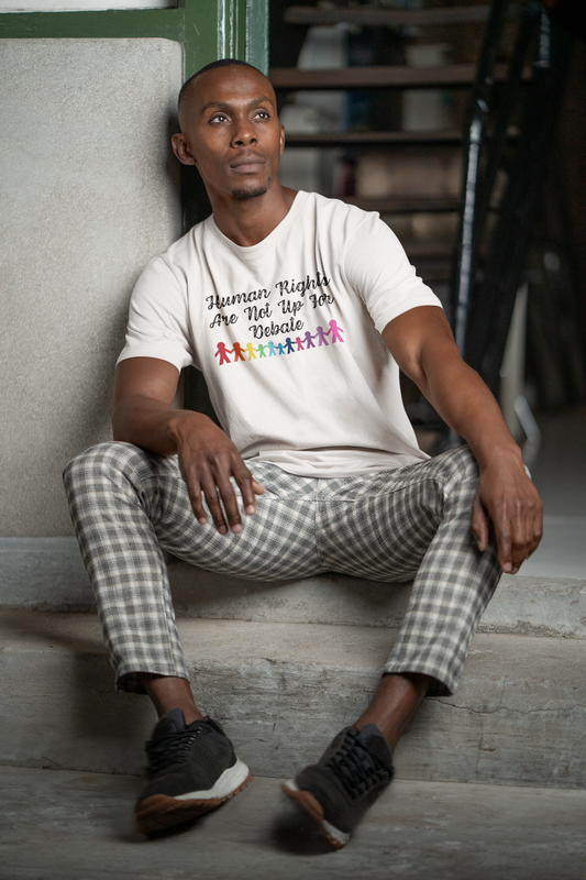 A man sits on concrete steps wearing a Classic White “Human Rights Are Not Up For Debate” t-shirt with rainbow figures, from The Unfussy Thread Collective.