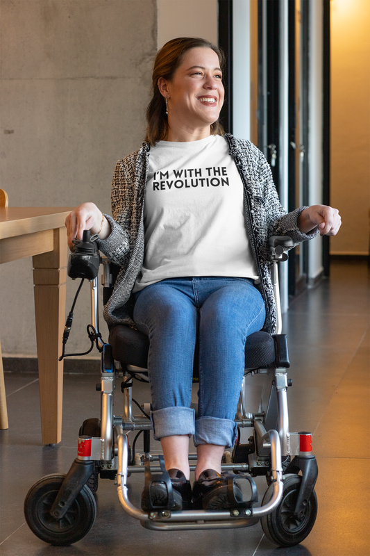 Smiling woman in a power wheelchair wearing a white “I’m With the Revolution” t-shirt, styled with jeans and a textured cardigan in a hallway setting.
