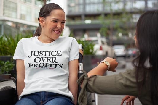 Smiling woman in a wheelchair wears a white “Justice Over Profits” t-shirt while bumping fists with a friend outdoors in an urban setting. She looks joyful and confident, embodying the shirt’s bold message advocating for equity over greed.