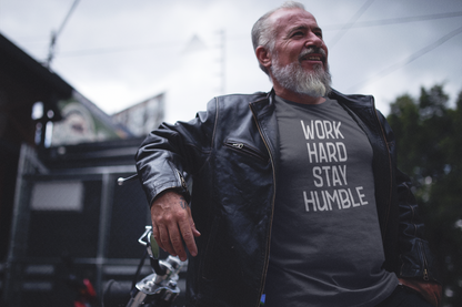 Older man in a leather jacket standing confidently next to a motorcycle, wearing a charcoal t-shirt that says "Work Hard Stay Humble" in bold lettering.