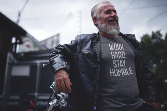 Older man in a leather jacket standing confidently next to a motorcycle, wearing a charcoal t-shirt that says "Work Hard Stay Humble" in bold lettering.