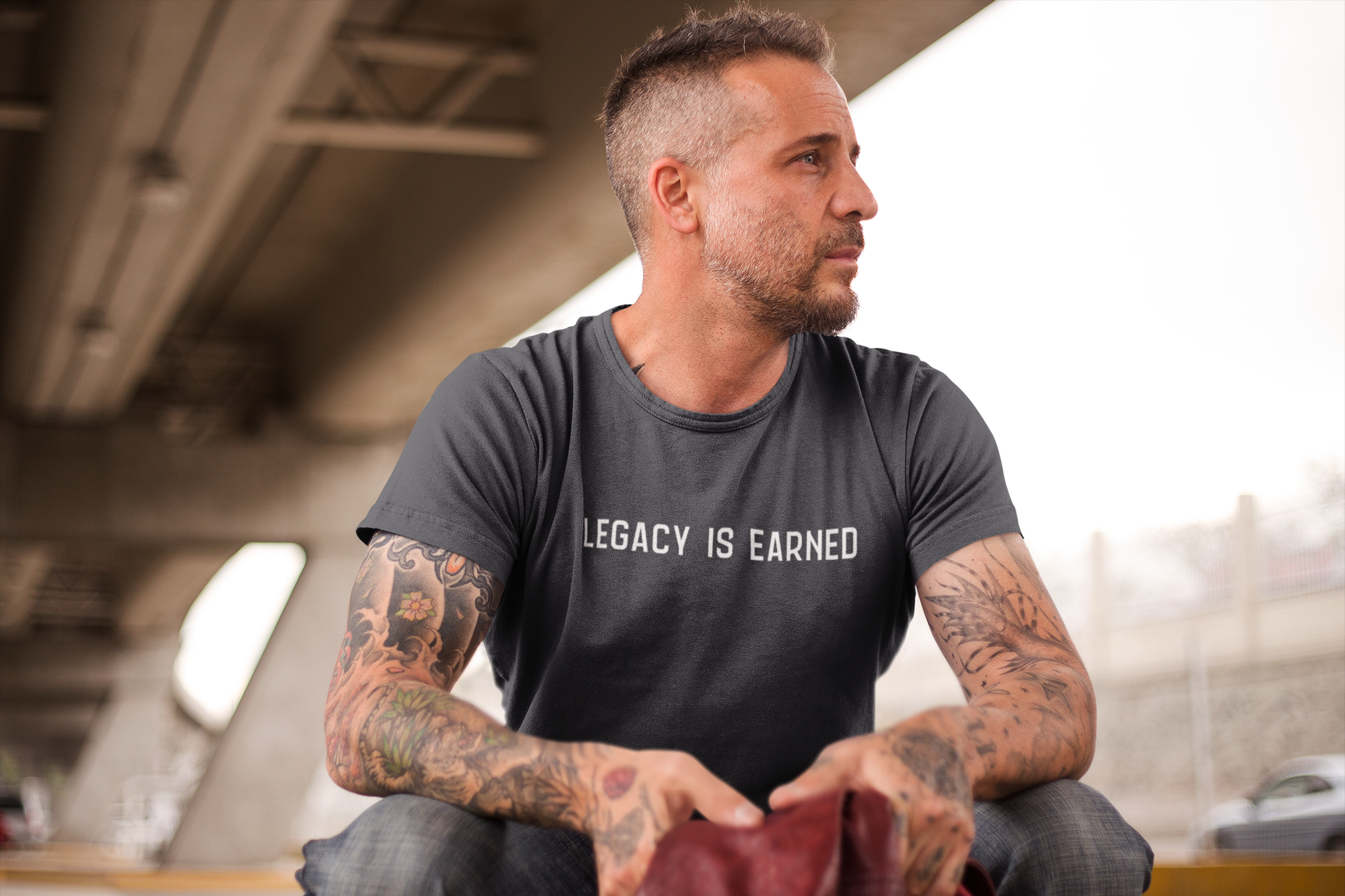 Middle-aged man with tattoos and a trimmed beard wearing a charcoal t-shirt that reads "Legacy Is Earned" while seated under an overpass.