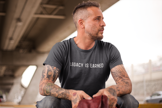 Middle-aged man with tattoos and a trimmed beard wearing a charcoal t-shirt that reads "Legacy Is Earned" while seated under an overpass.
