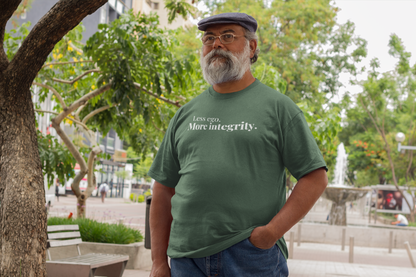 Older man with a gray beard and glasses standing outdoors near a fountain, wearing a green “Less ego. More integrity.” t-shirt and a newsboy cap.