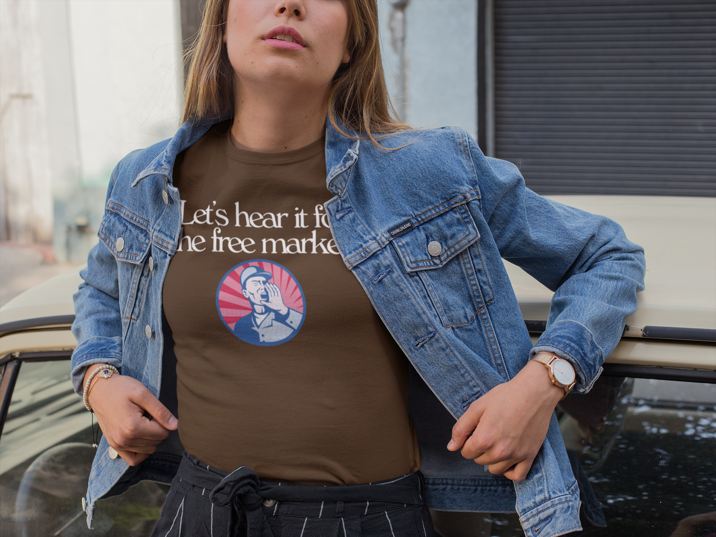 A woman wearing a brown t-shirt with the phrase “Let’s hear it for the free market” and a vintage-style illustration of a man shouting stands confidently next to a classic car. She wears a denim jacket and black striped pants, evoking a casual, bold, and slightly retro vibe.