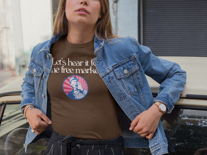 A woman wearing a brown t-shirt with the phrase “Let’s hear it for the free market” and a vintage-style illustration of a man shouting stands confidently next to a classic car. She wears a denim jacket and black striped pants, evoking a casual, bold, and slightly retro vibe.