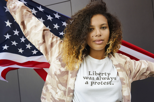 A woman stands confidently in front of a U.S. flag wearing a white t-shirt that reads “Liberty was always a protest.” She wears a beige camo jacket and gazes ahead with calm strength.