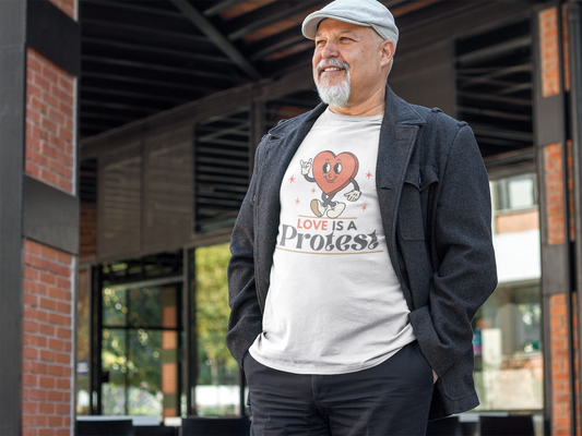An older man with a gray beard and a flat cap smiles warmly while standing outside a cafe. He wears a white t-shirt featuring a vintage cartoon heart with sneakers and gloves, surrounded by small stars, and the words “Love Is a Protest” in bold, retro-style lettering.