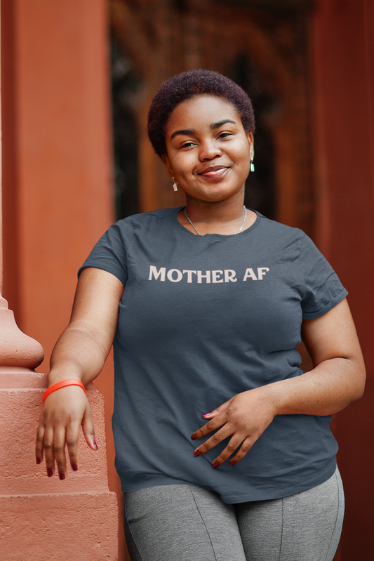 Smiling woman with short afro hairstyle wearing a blue "Mother AF" t-shirt from The Unfussy Thread Collective, leaning on a peach column outdoors.