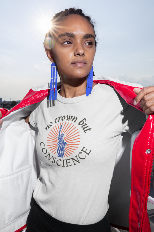 A woman in bold blue earrings and a red jacket opens her coat to reveal a white t-shirt that reads “no crown but CONSCIENCE” with a vintage Statue of Liberty design. The sun flares behind her as she looks into the distance with confidence.