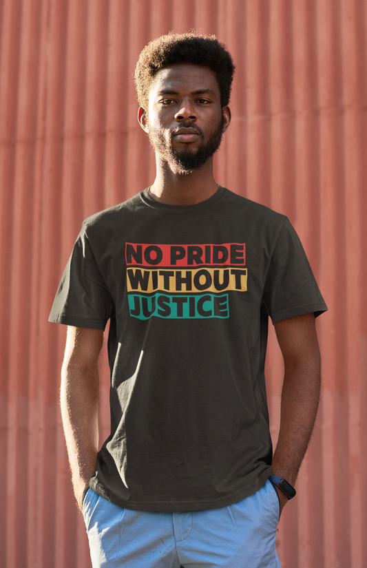 A serious young Black man stands in front of a textured, rust-red wall wearing a dark t-shirt that reads “No Pride Without Justice” in red, gold, and green block letters. His hands are in his pockets and the sunlight casts strong shadows, emphasizing the shirt’s powerful call for intersectional justice and racial equity within the LGBTQIA+ movement.