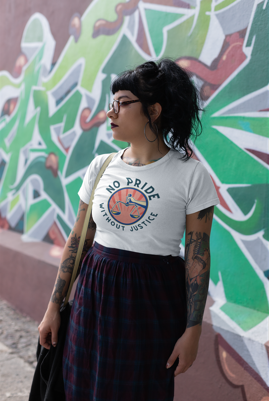 A young woman with tattoos and hoop earrings stands confidently in front of a graffiti-covered wall, wearing a white t-shirt with a retro-style scale of justice and the words “No Pride Without Justice” in bold lettering. She looks off to the side with purpose, her black bangs and plaid skirt adding to the rebellious yet grounded vibe of the shot.