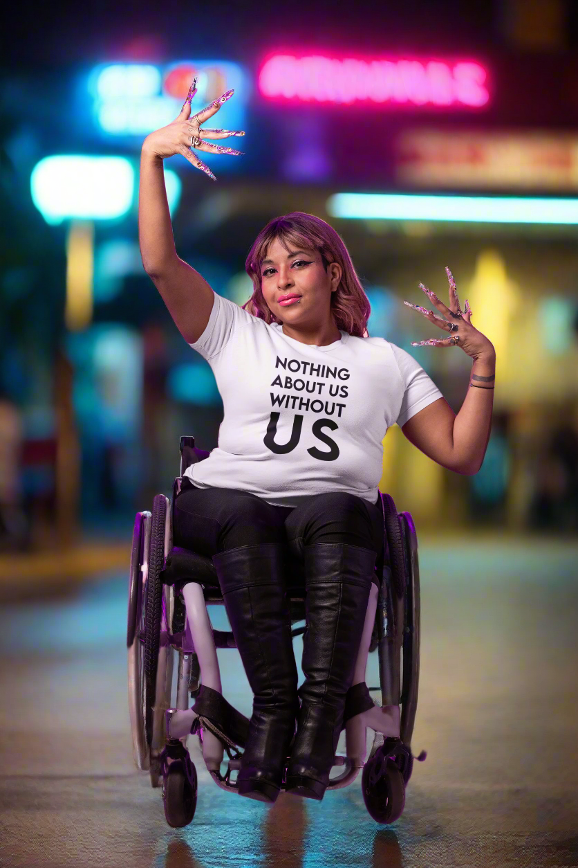 A stylish woman in a wheelchair strikes a powerful pose at night, lit by neon signs in the background. She wears thigh-high boots and a bold white t-shirt that reads “Nothing About Us Without Us.” Her long, decorative nails and confident expression emphasize unapologetic pride, visibility, and disabled empowerment.