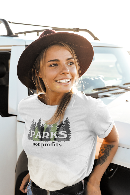 Smiling woman in a wide-brim hat leans against a white SUV, wearing a “Parks Not Profits” t-shirt featuring a forest graphic, embodying eco-conscious values and public land protection.