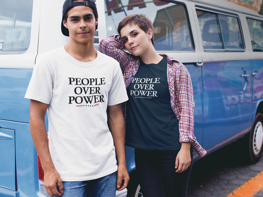 Two young adults wearing “People Over Power” t-shirts stand in front of a blue retro van. The person on the left wears a white tee with the slogan in bold black letters accented by small stars and stripes. The person on the right leans casually, wearing a black version of the same shirt layered under an open plaid button-up. Both exude a casual, activist vibe.