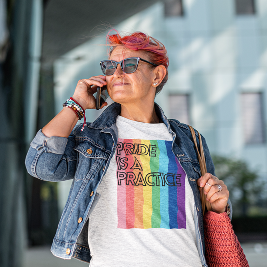 A confident older woman with pink hair stands outside a modern building, talking on her phone. She wears a heather gray t-shirt that says “Pride Is a Practice” over bold rainbow stripes. Paired with a denim jacket, sunglasses, and a coral tote, the look radiates queer joy and seasoned self-assurance.