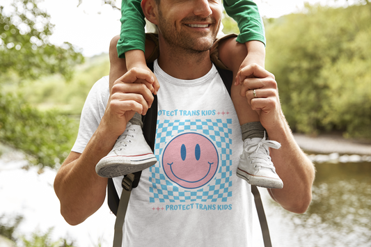 A man smiles while carrying a child on his shoulders during a nature walk. He’s wearing a white t-shirt that reads “Protect Trans Kids” above and below a large pink smiley face, centered on a blue-and-white checkerboard pattern. The design blends retro Y2K vibes with a bold, affirming message of trans allyship.