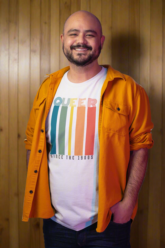 A smiling man in a mustard-yellow overshirt stands confidently against a wood-paneled background. His white t-shirt features the phrase “Queer Since the 1900s” in bold vintage typography, with vertical rainbow stripes in muted retro tones. His expression radiates pride, comfort, and a sense of identity.