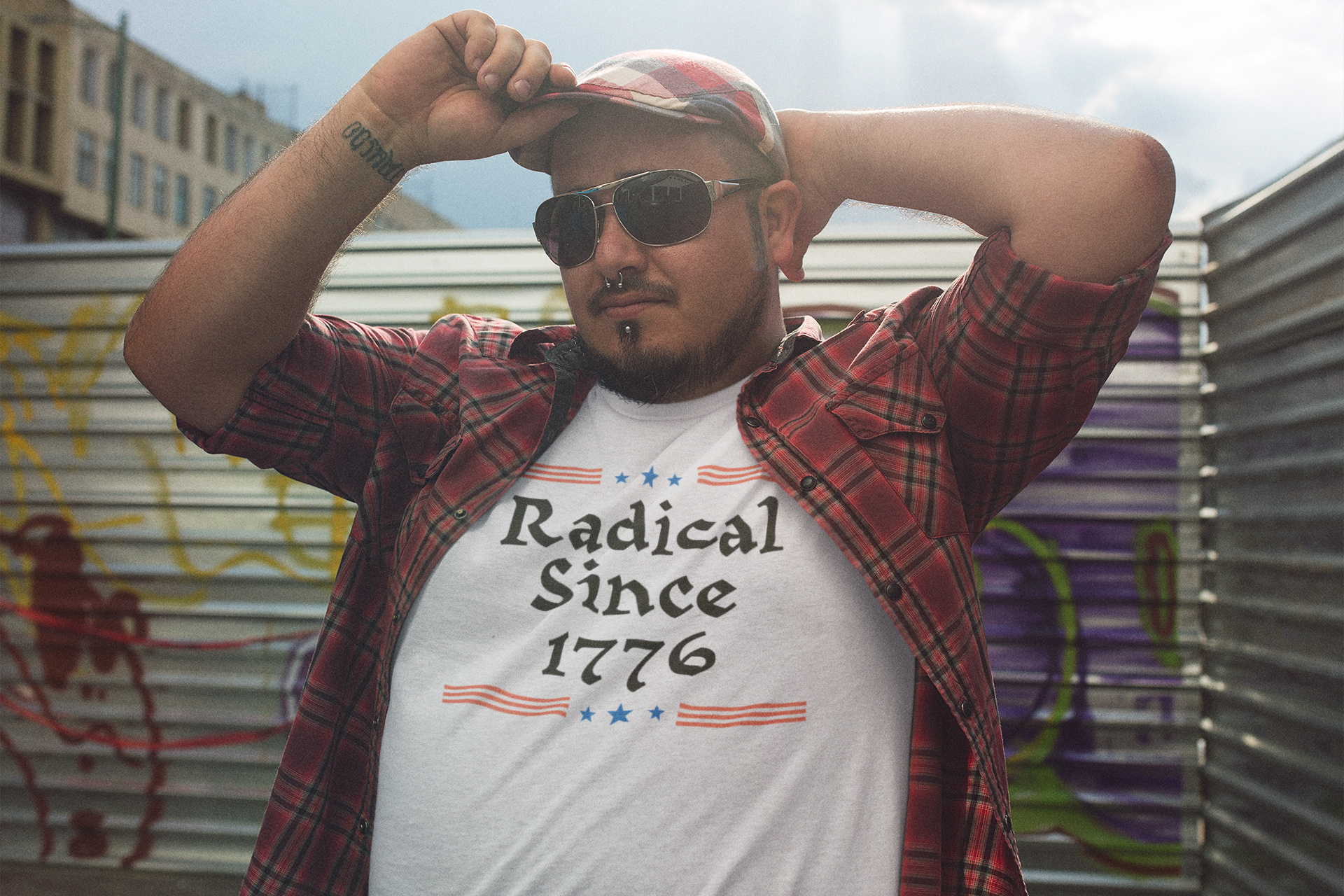 A man in sunglasses and a red flannel shirt poses confidently in an urban alleyway, wearing a white t-shirt that reads “Radical Since 1776” with red and blue graphics.