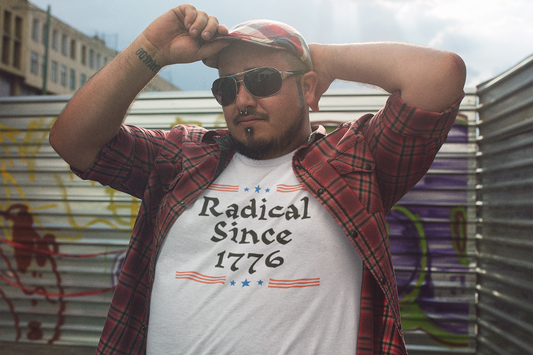 A man in sunglasses and a red flannel shirt poses confidently in an urban alleyway, wearing a white t-shirt that reads “Radical Since 1776” with red and blue graphics.