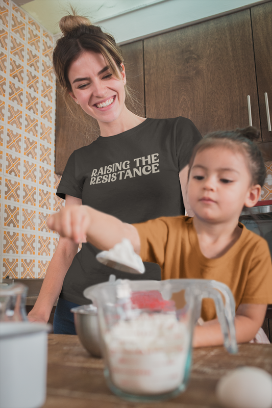 A smiling mother wears a “Raising the Resistance” t-shirt in the kitchen while baking with her young daughter, from The Unfussy Thread Collective.