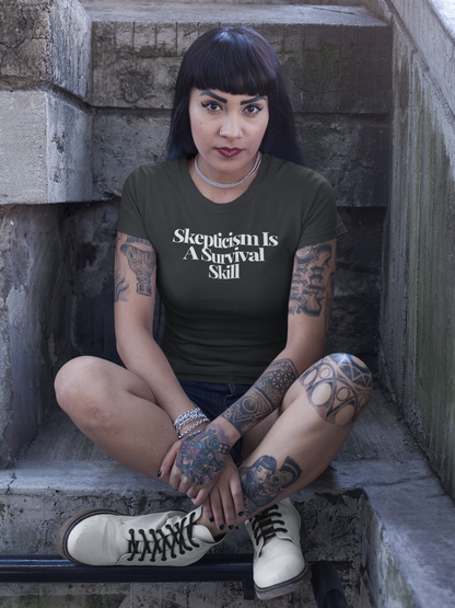 Tattooed woman with dark hair and bangs sits cross-legged on a stone stairway, wearing a dark “Skepticism Is A Survival Skill” t-shirt. Her intense gaze and bold style amplify the shirt’s message about resilience and critical thinking.