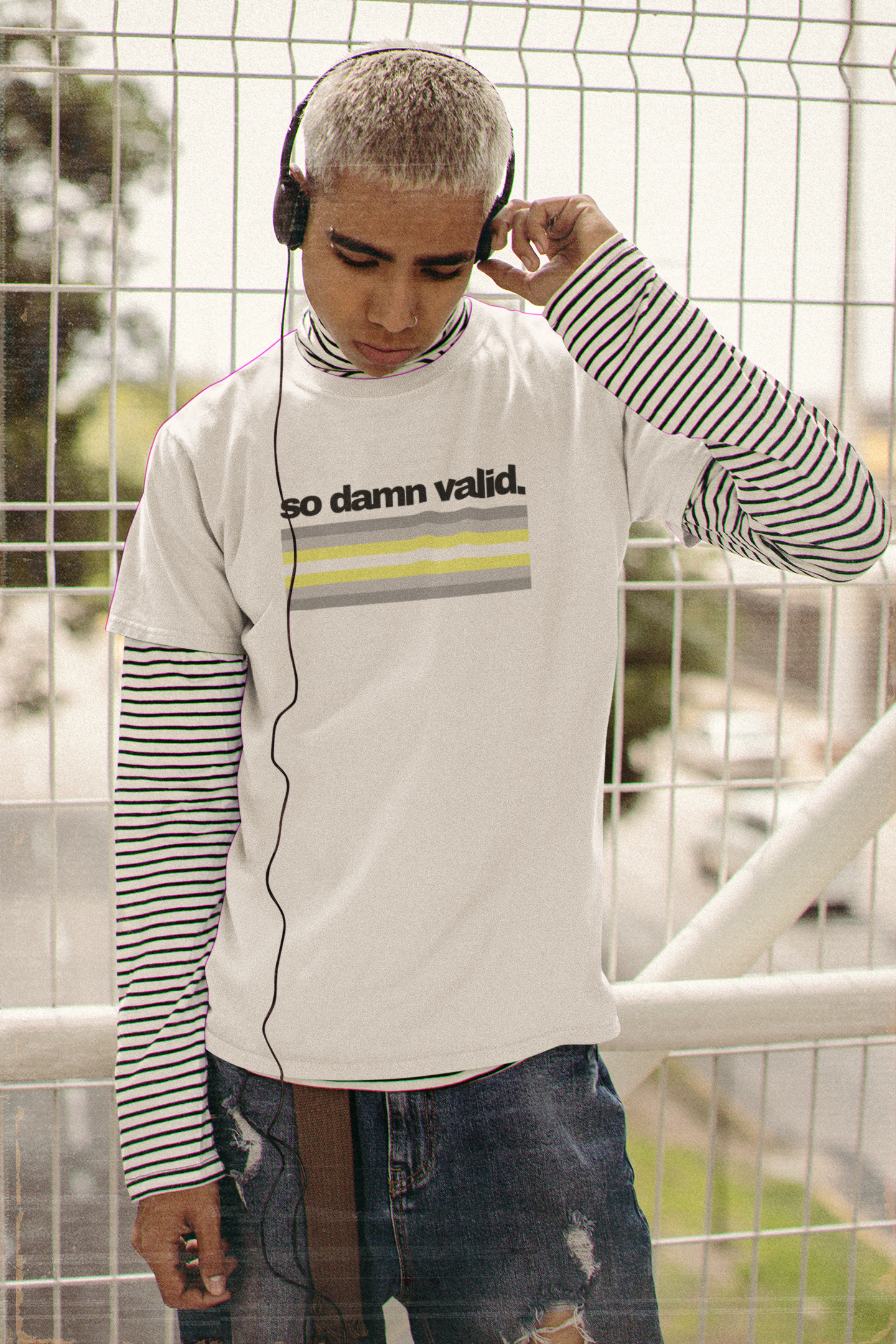 Person wearing a white t-shirt with the phrase "so damn valid." above horizontal demigender pride stripes in black, gray, yellow, and white, layered over a striped long sleeve and paired with ripped jeans.