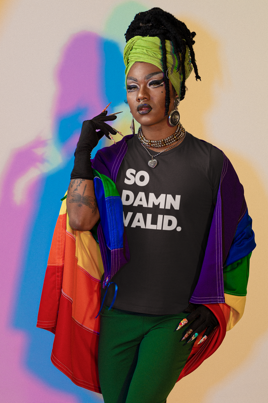 A bold, expressive person in a rainbow cape and green head wrap poses confidently against a colorful shadow backdrop, wearing a black t-shirt that reads “So Damn Valid.”
