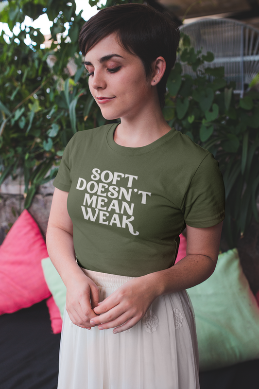 A woman with short hair wears a Heather Olive “Soft Doesn’t Mean Weak” t-shirt while standing calmly in a cozy outdoor setting, part of The Unfussy Thread Collective.