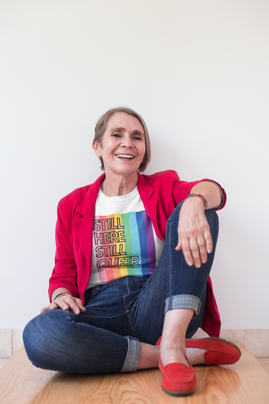 A joyful older woman in a red blazer and jeans sits casually on a wooden bench, smiling confidently. She wears a white t-shirt that reads “Still Here Still Queer” over a rainbow-striped background, proudly celebrating LGBTQ+ identity.