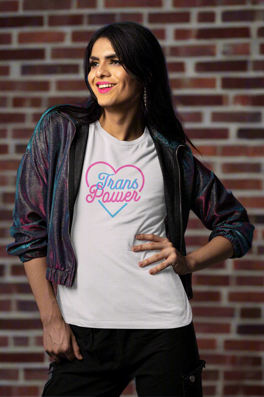A confident woman smiles brightly while wearing a white “Trans Power” t-shirt featuring pink and blue lettering inside a heart outline. She stands against a brick wall backdrop, exuding pride and strength in a shimmering jacket.