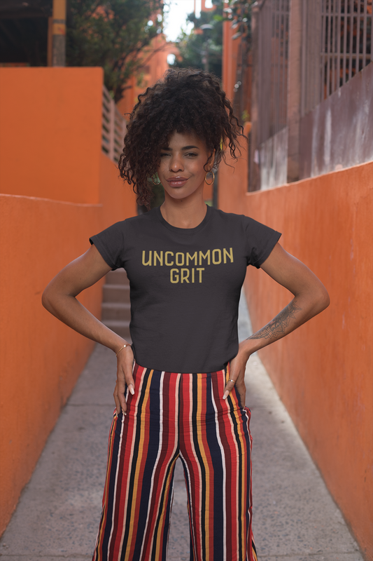 A confident woman with curly hair stands in a vibrant orange alleyway, wearing a black "Uncommon Grit" t-shirt and striped pants, hands on hips and smiling slightly.