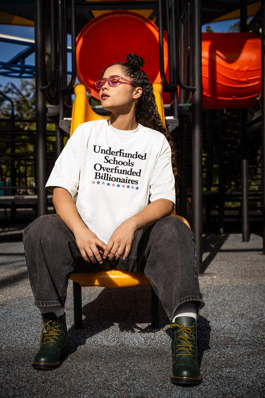 A woman wearing an oversized white t-shirt with the slogan “Underfunded Schools Overfunded Billionaires” sits confidently on a yellow playground slide. She wears dark jeans, green boots with yellow laces, and pink sunglasses. The playground equipment in the background contrasts with the serious message on her shirt.