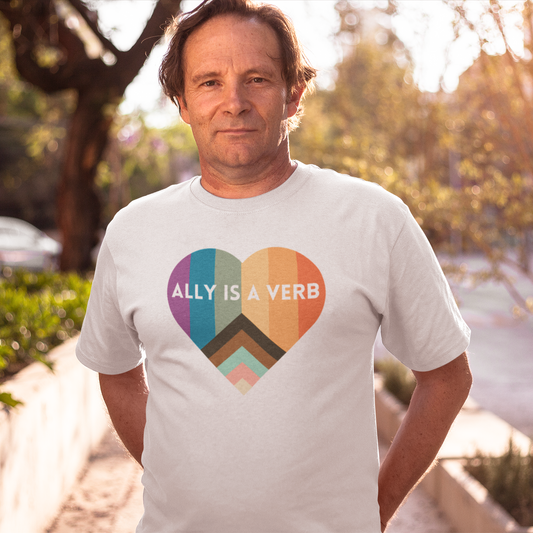 A middle-aged man stands outdoors at golden hour wearing a white t-shirt with a rainbow-striped heart that reads “Ally Is a Verb,” promoting LGBTQ+ allyship.