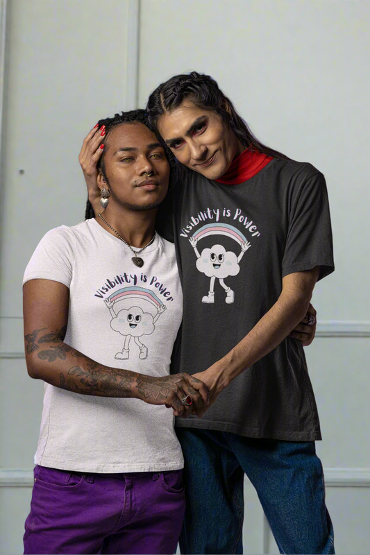 Two people embrace warmly against a vibrant blue and pink background, both wearing “Visibility is Power” t-shirts featuring a joyful cloud and rainbow design. Their closeness and expressions radiate pride and authenticity.
